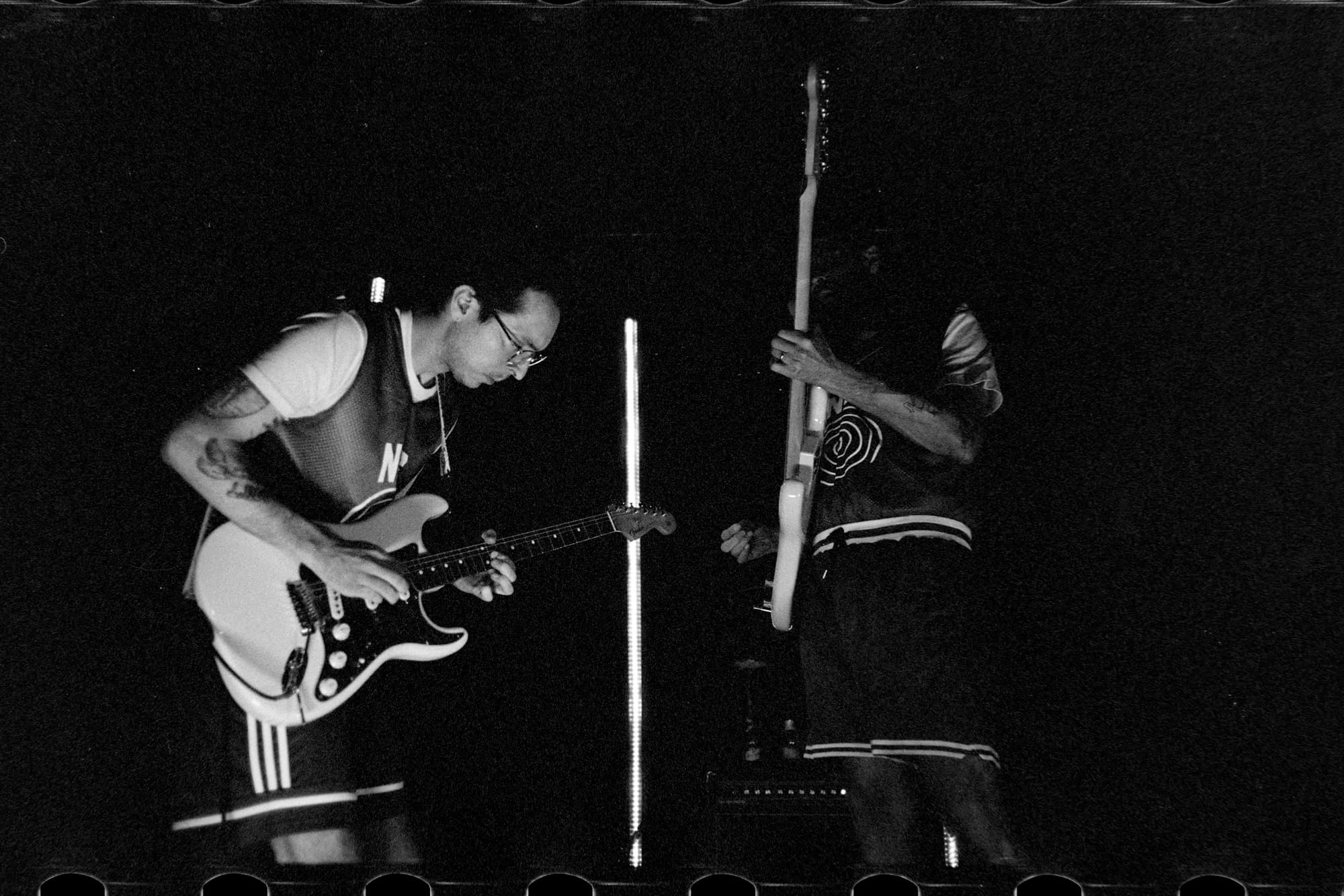 2 guys playing guitars on a very dark stage and they are the only to bright things visible.