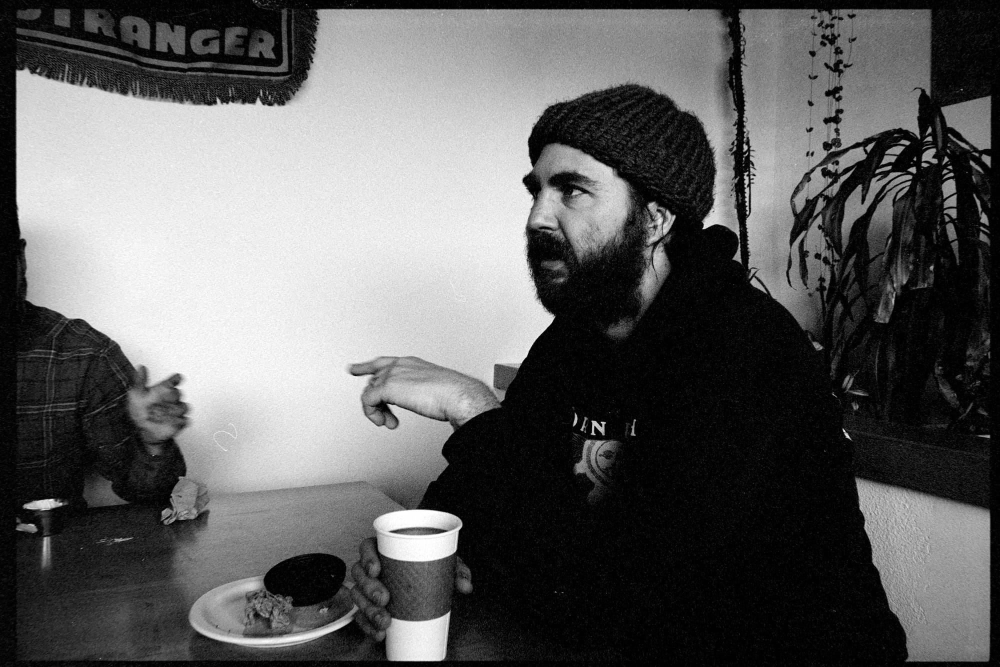 A black and white photo of a guy talking while holding a coffee and has a plate with a bagel on it.