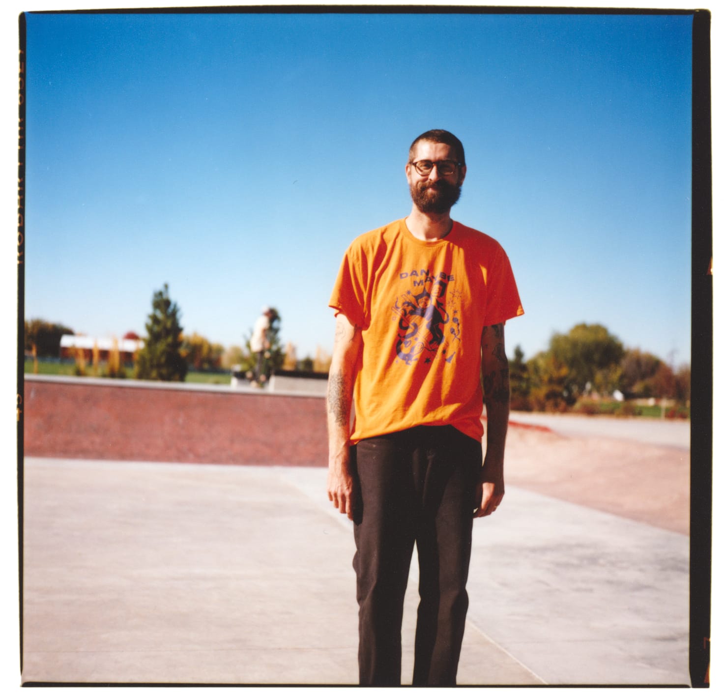 1 guy standing in an empty skatepark with red ramps in the background and a blue sky.