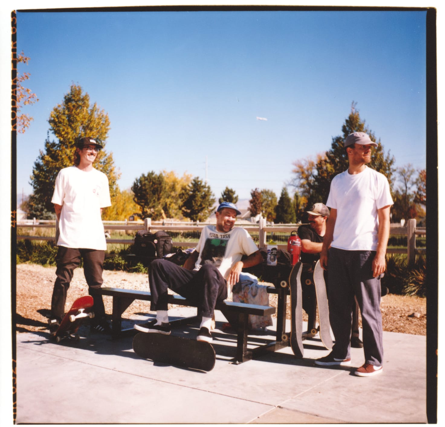 4 guys sitting and standing around a picnic table on a bright sunny day.